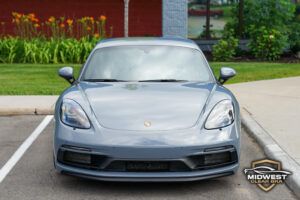 Gray Porsche Cayman after professional paint protection film (PPF), ceramic coating, and window tint installation, shown at a front three quarter angle with clear bra film protecting the hood, bumper, and fenders.